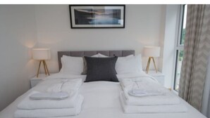 A neatly made bed with white linens and two pillows, a gray accent pillow, and two bedside lamps with wooden bases. - Tailored Stays  -  Executive Town House (Cambridge)