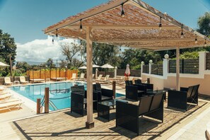 A poolside area with lounge chairs and umbrellas, a wooden deck, and a pergola with hanging lights. - Kalypso Studios (Corfu)