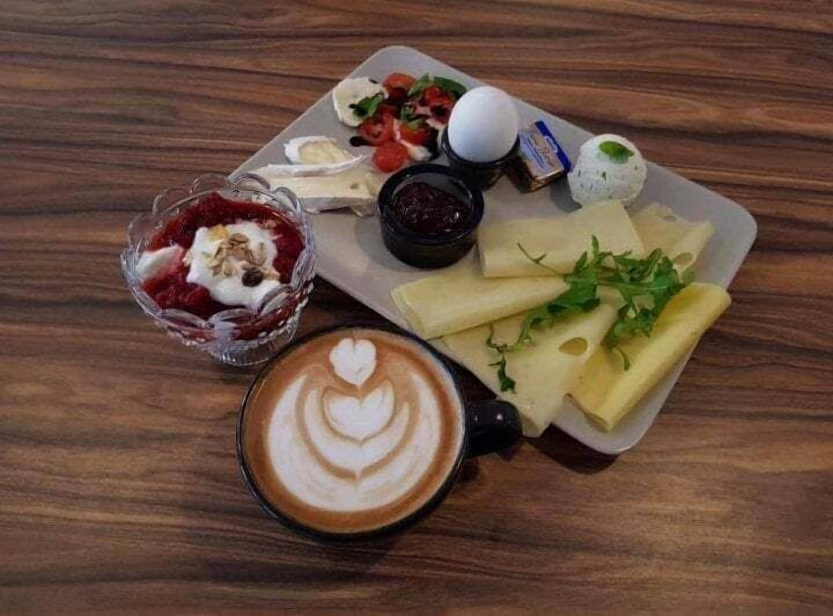 A breakfast setting with a plate of cheese, a glass of red drink, a cup of coffee with latte art, and a small bowl of jam.