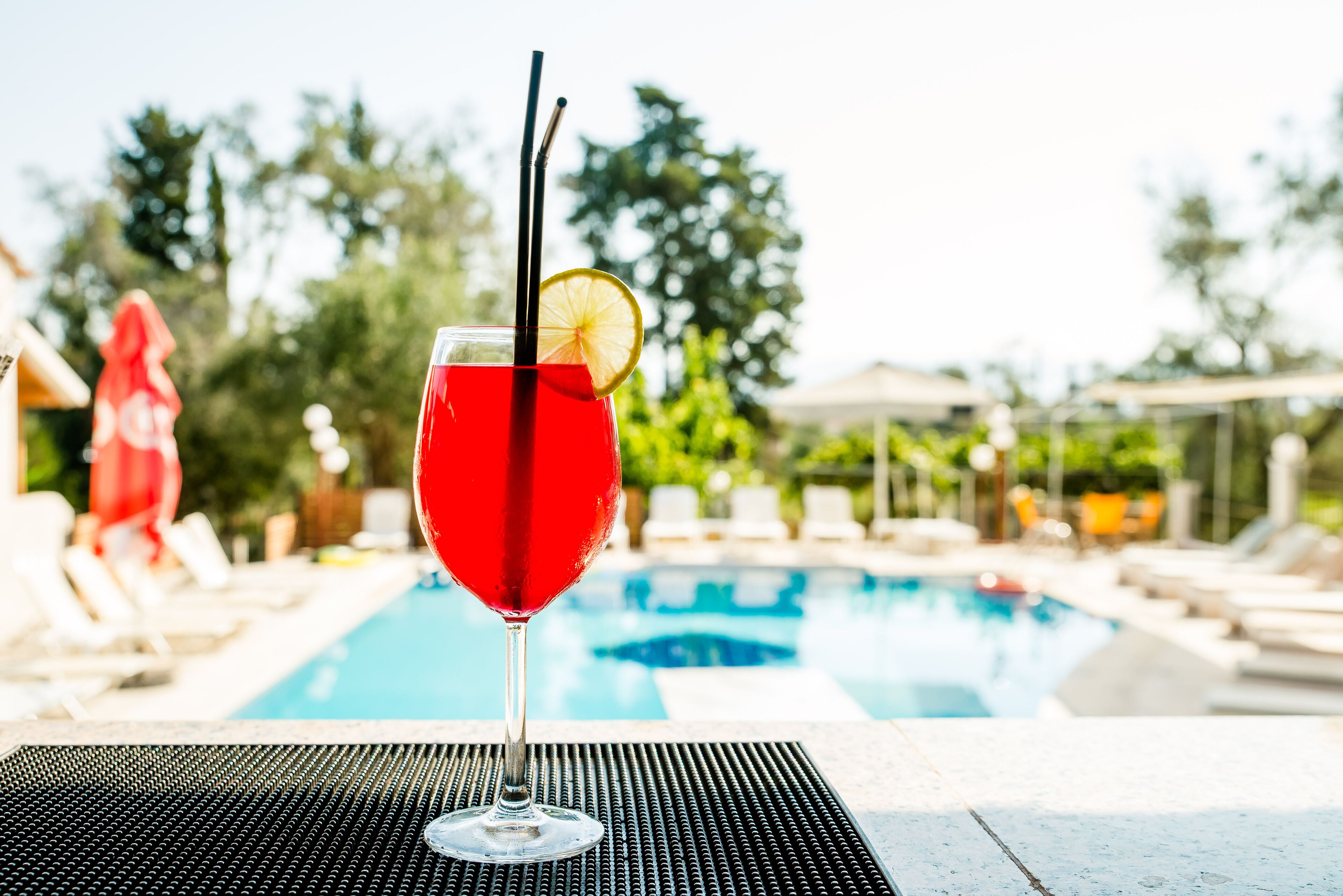Un cocktail rouge, garni d’une tranche de citron et accompagné de pailles, est posé sur une table au bord d’une piscine.