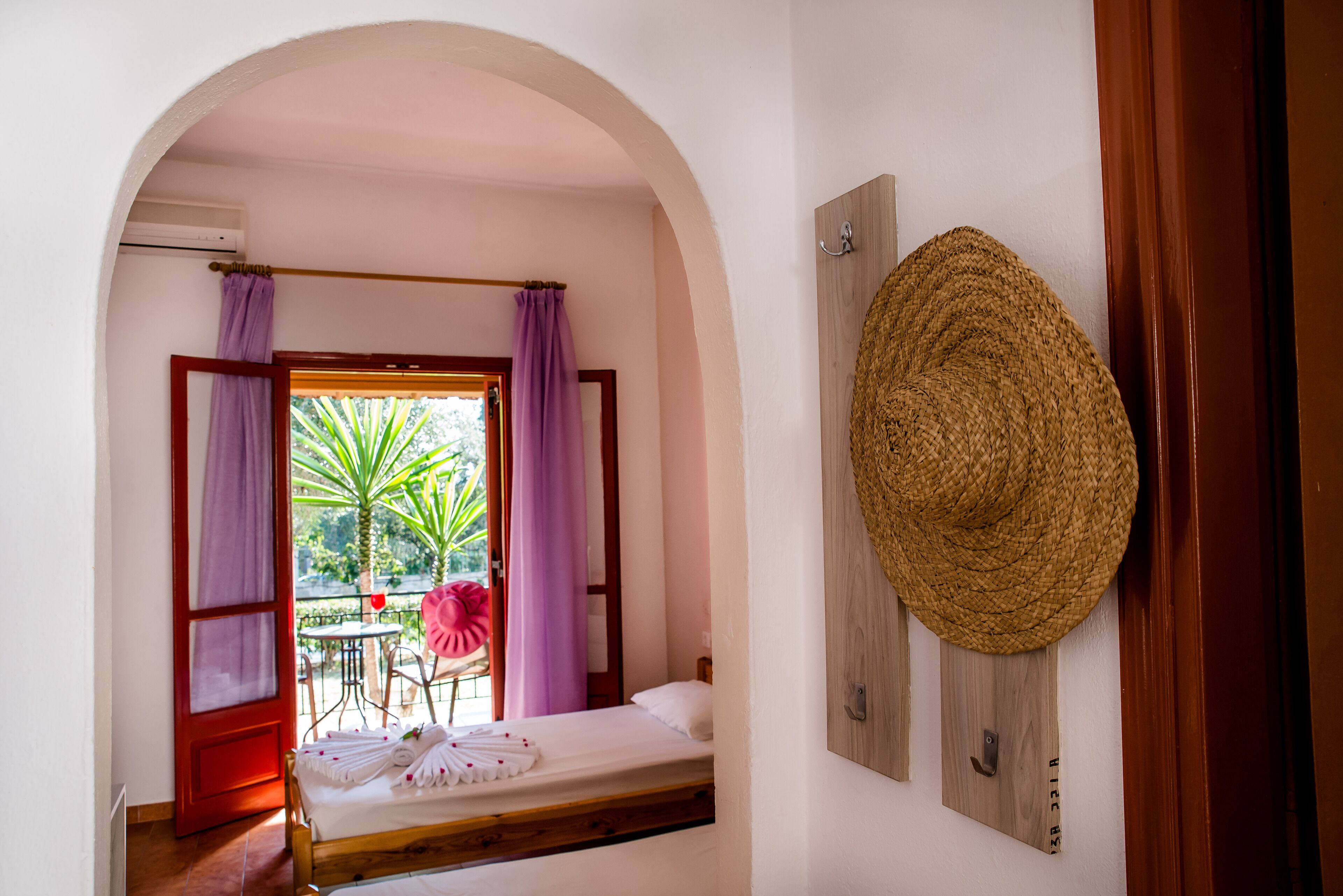 A room with a bed, a window with purple curtains, a straw hat on the wall, and a small table with a chair outside.