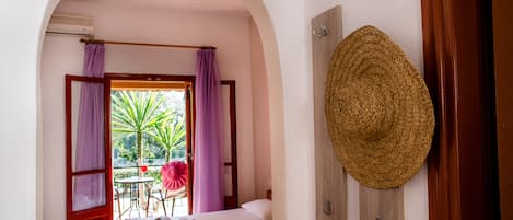 A room with a bed, a window with purple curtains, a straw hat on the wall, and a small table with a chair outside.