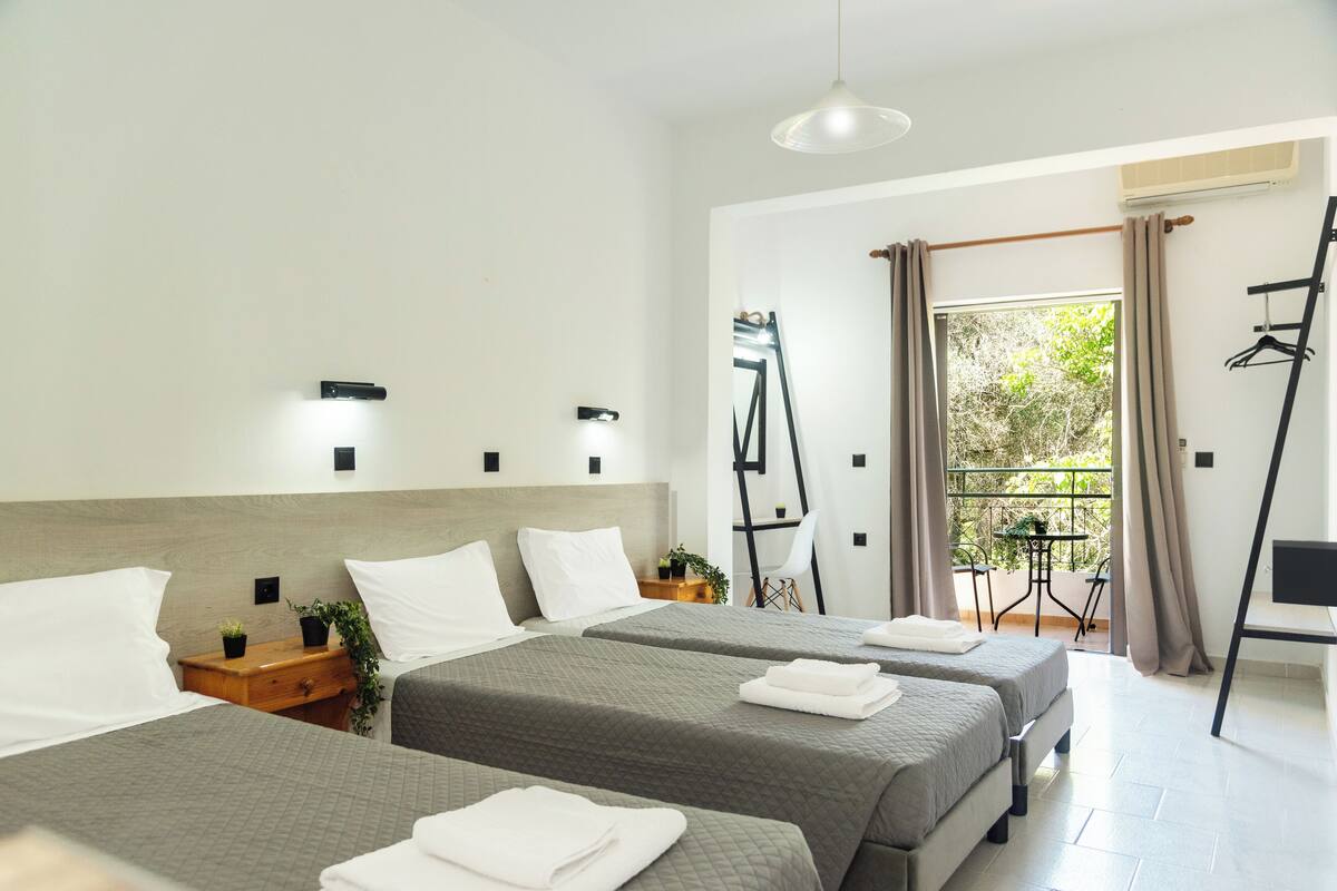 Habitación de hotel con dos camas, una mesita, una silla, una planta y un balcón con cortinas.