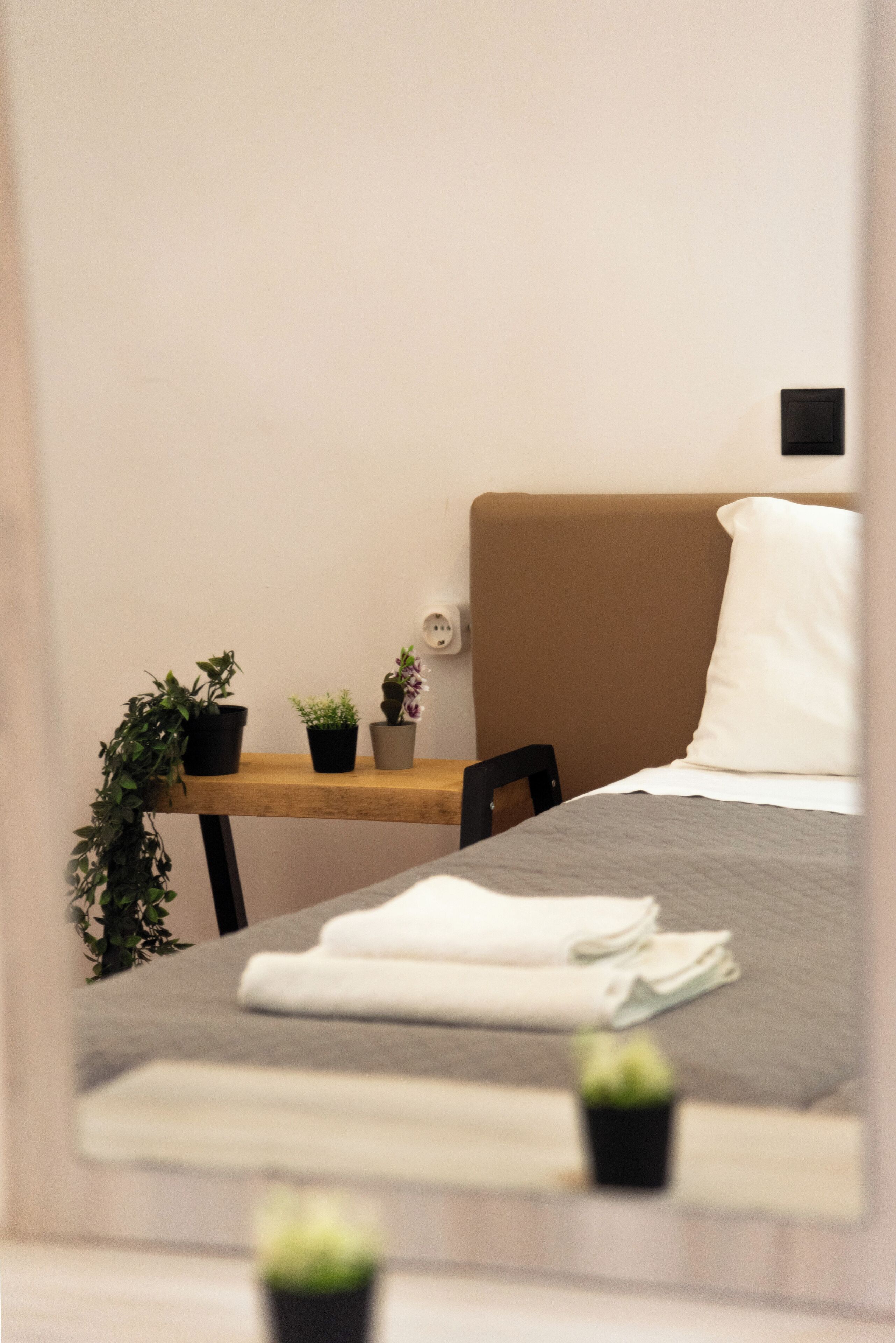 A neatly made bed with a grey blanket and white pillows, a small wooden table with potted plants, and a black chair.