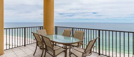 A balcony with a glass table and chairs, overlooking a beach and ocean.