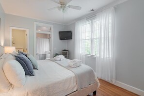 Une chambre à coucher avec un lit, un ventilateur de plafond, une télévision et une fenêtre avec des rideaux.