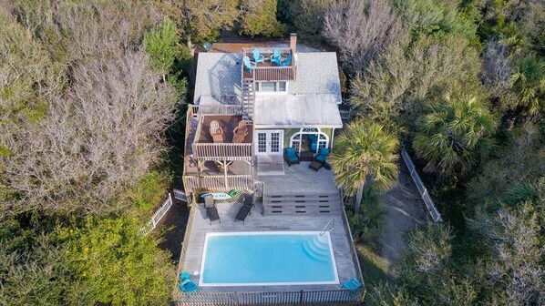 Vue aérienne d’une maison avec une piscine, des chaises longues et une terrasse clôturée, le tout entouré d’arbres.