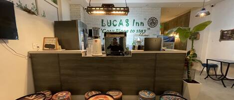 A reception area with a sign reading "LUCAS INN" and a slogan "Come to stay". There are four stools with a coffee theme, a plant, and a TV mounted on the wall.