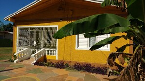 Une maison jaune avec une fenêtre blanche et une porte grillagée, un escalier et un jardin où poussent des plantes violettes.