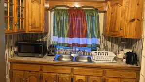 Une cuisine avec des meubles de rangement en bois, un four à micro-ondes, un grille-pain, un évier et une fenêtre agrémentée de rideaux colorés.