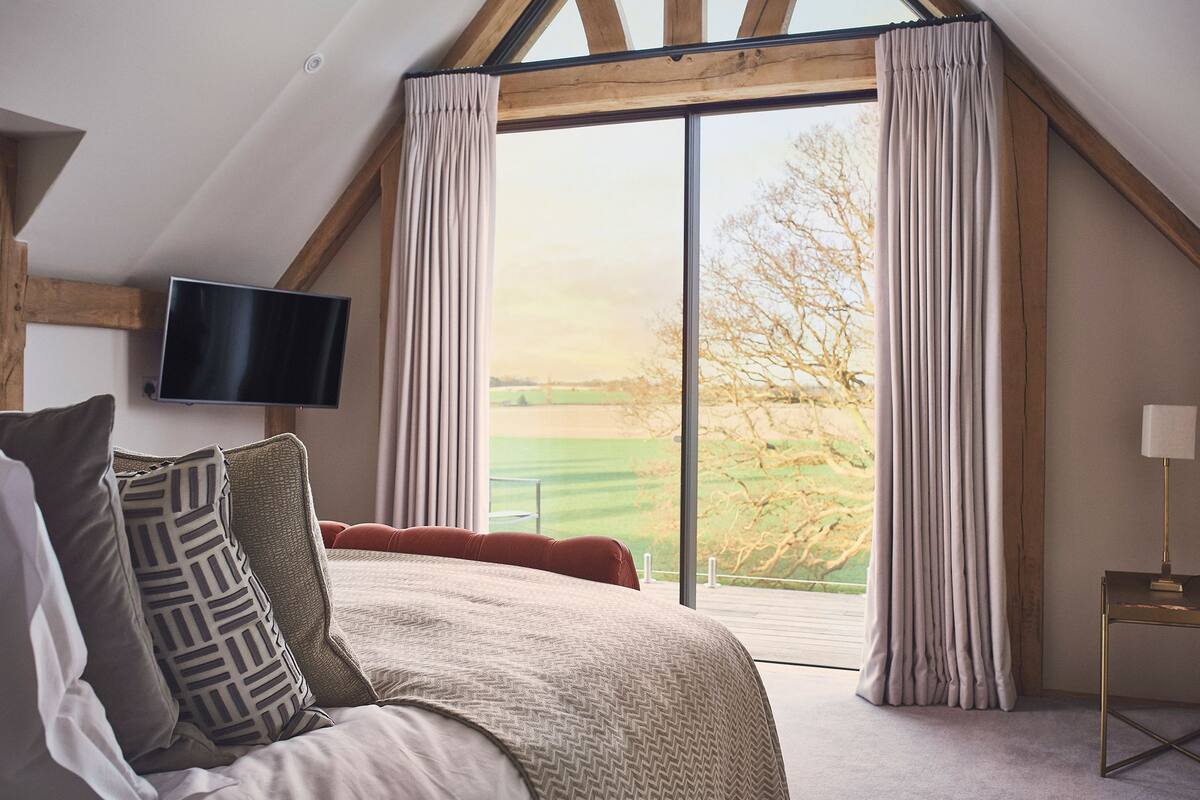 A bedroom with a large bed, a flat-screen TV mounted on the wall, and a view of a green landscape through a large window.