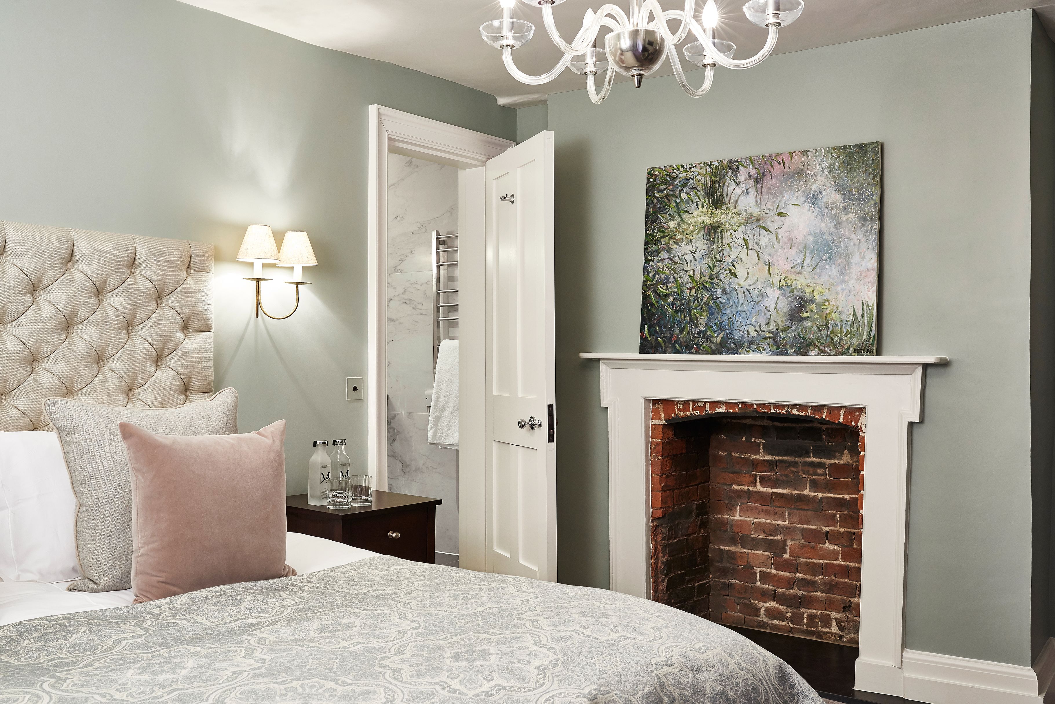 A bedroom with a tufted headboard, a bed with patterned bedding, a nightstand with a lamp, a white door, a fireplace, and a painting on the wall.