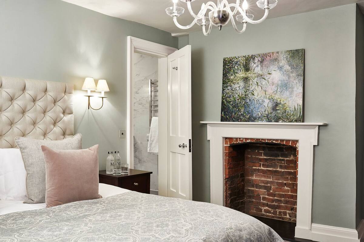 A bedroom with a tufted headboard, a bed with patterned bedding, a nightstand with lamps, a fireplace, and a painting on the wall.