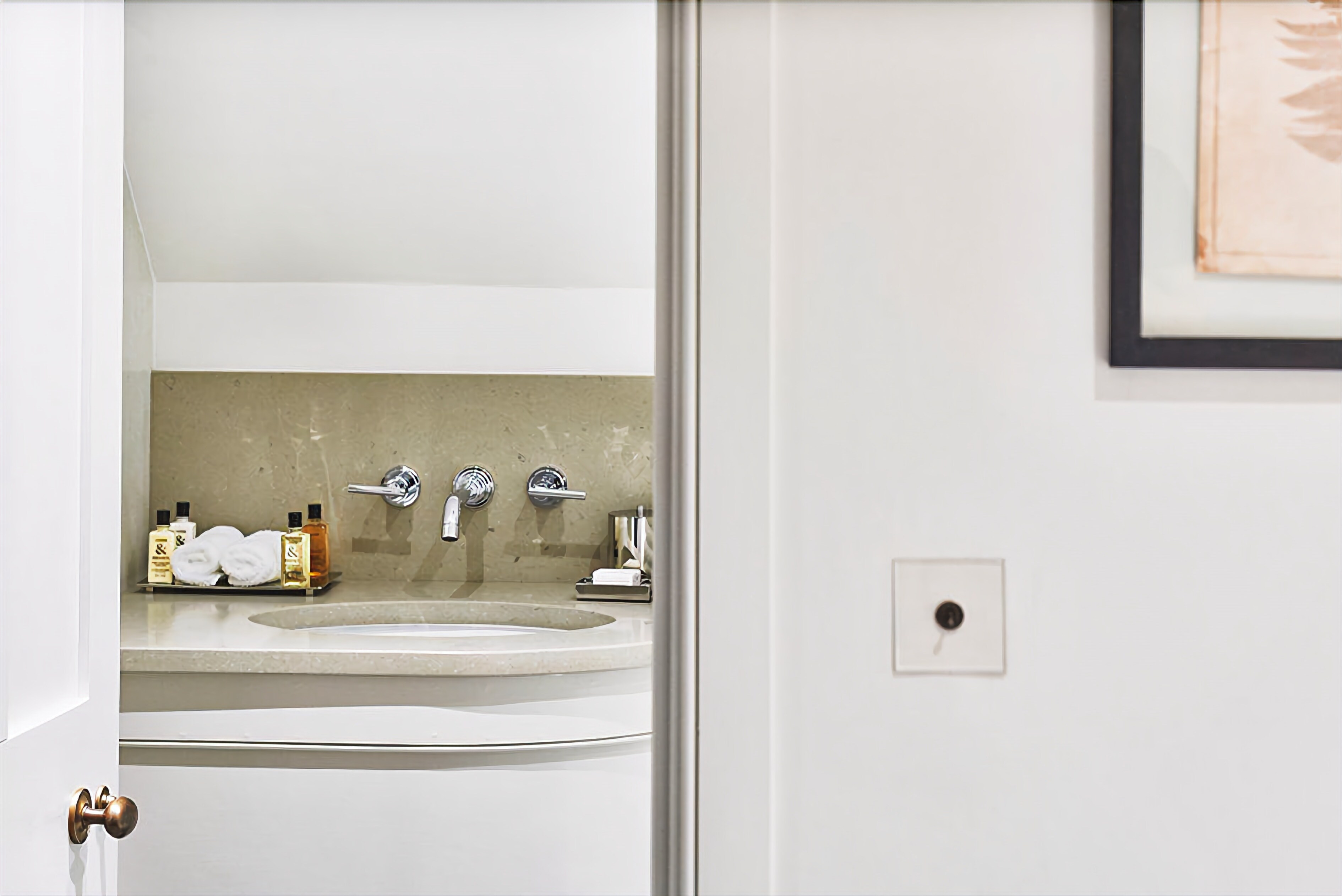 Un baño con un lavamanos blanco, dos grifos y un salpicadero de azulejos color crema.
