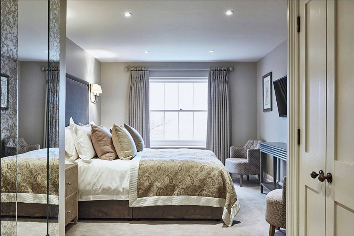 A bedroom with a large bed, a desk, and a chair. There is a window with curtains, a wall-mounted lamp, and a framed picture.