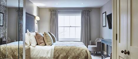 A bedroom with a large bed, a desk, and a chair. There is a window with curtains, a wall-mounted lamp, and a framed picture.