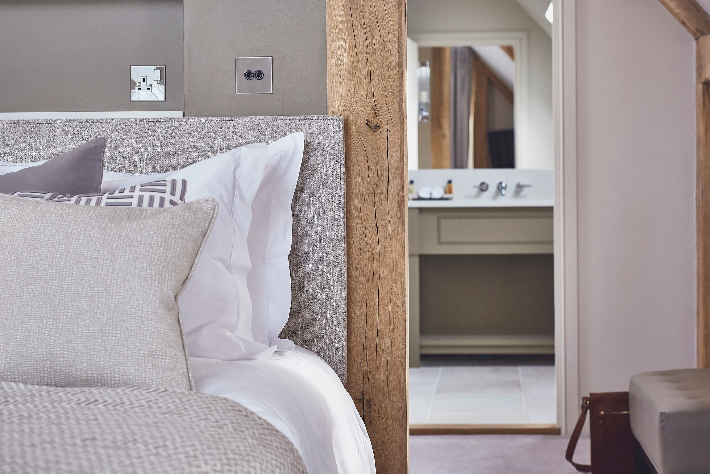 A neatly made bed with white and patterned pillows, a wooden headboard, and a view into a bathroom with a mirror and sink.