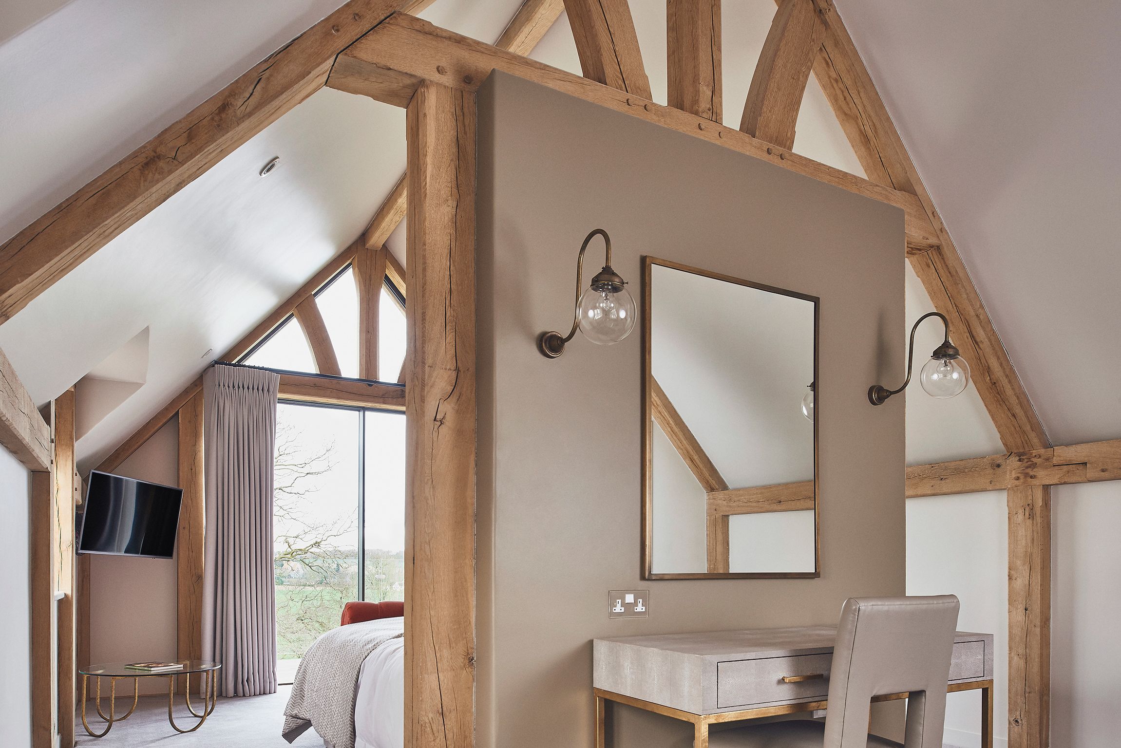 A bedroom with a large window, wooden beams, a bedside table, a chair, and a mirror.