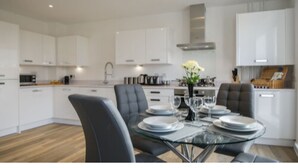 Una cocina moderna con armarios blancos, campana de acero inoxidable y un comedor con mesa de vidrio y sillas grises.