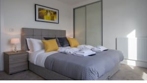 Un dormitorio moderno con una cama gris, almohadas blancas y amarillas, una mesita de noche con una lámpara y un ventanal grande.