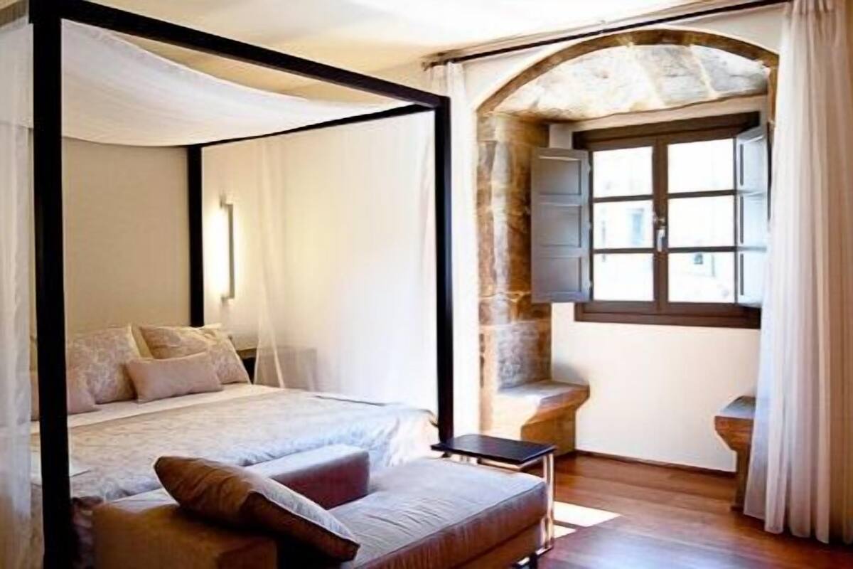 A bedroom with a four-poster bed, a small table, and a stone arch window.