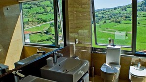 Un baño con lavamanos, inodoro y ventanas amplias que dan a campos verdes y un edificio lejano.