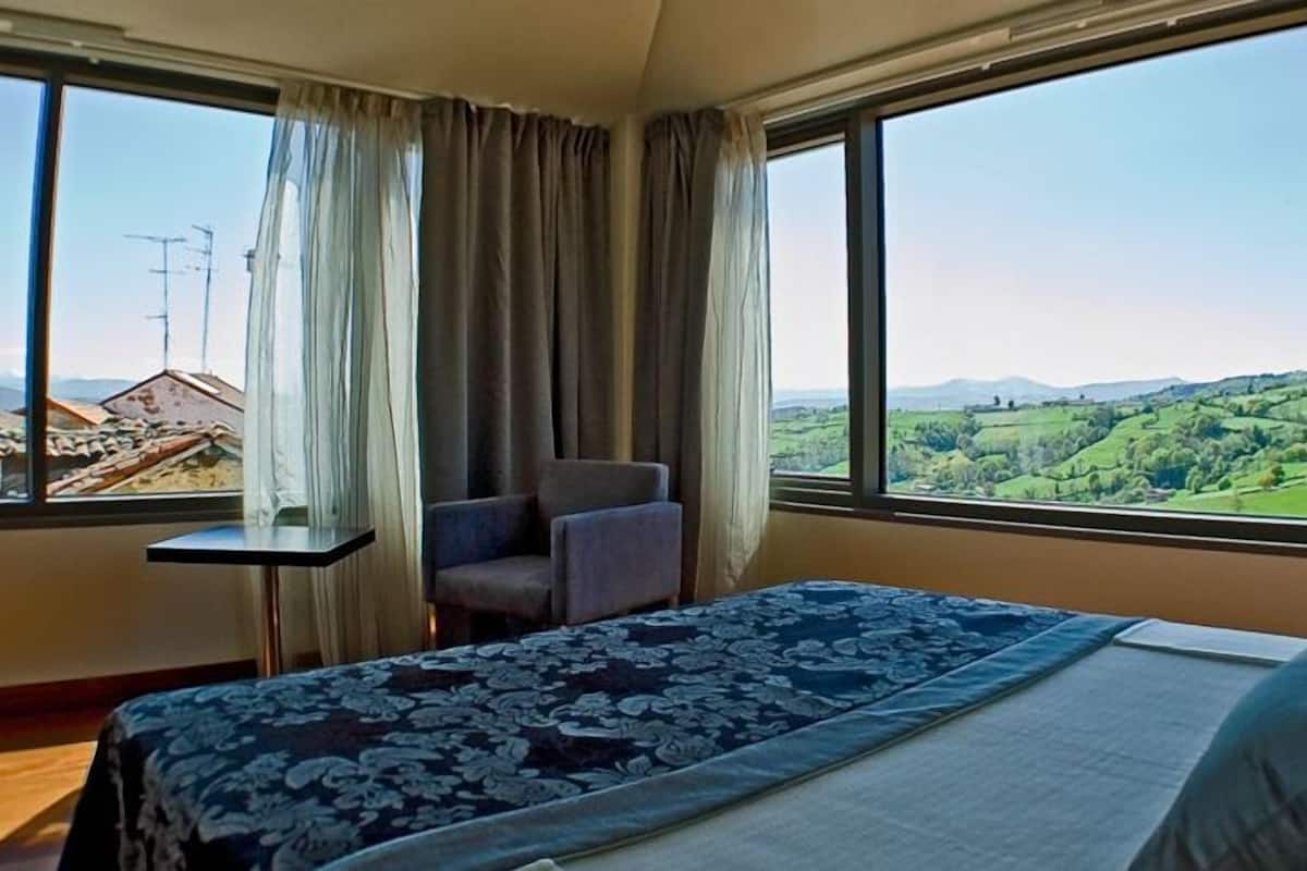 A hotel room with a bed, a chair, a small table, and large windows offering a view of a green landscape and distant buildings.