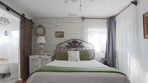 Un dormitorio con cama, mesita de noche, lámpara, un cuadro en la pared y ventana con cortinas.