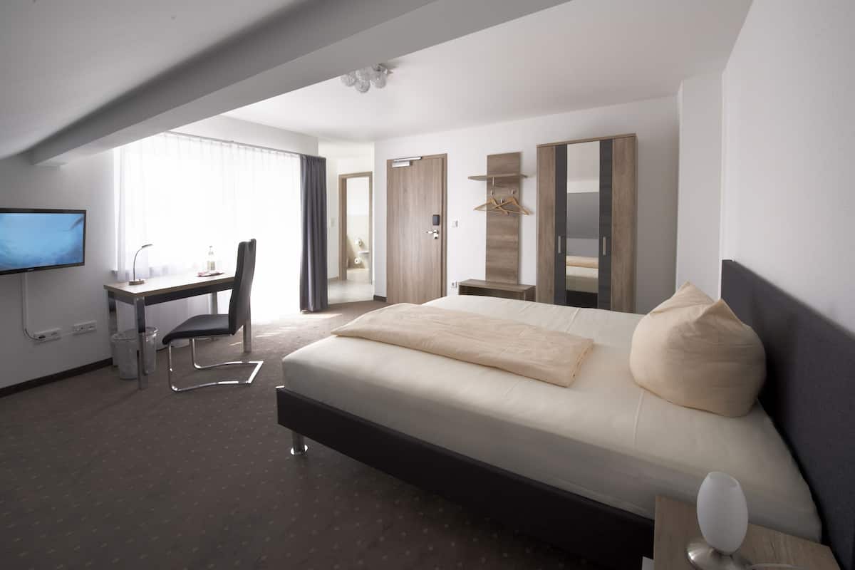 A modern hotel room with a large bed, a desk with a chair, a TV, and a wardrobe.