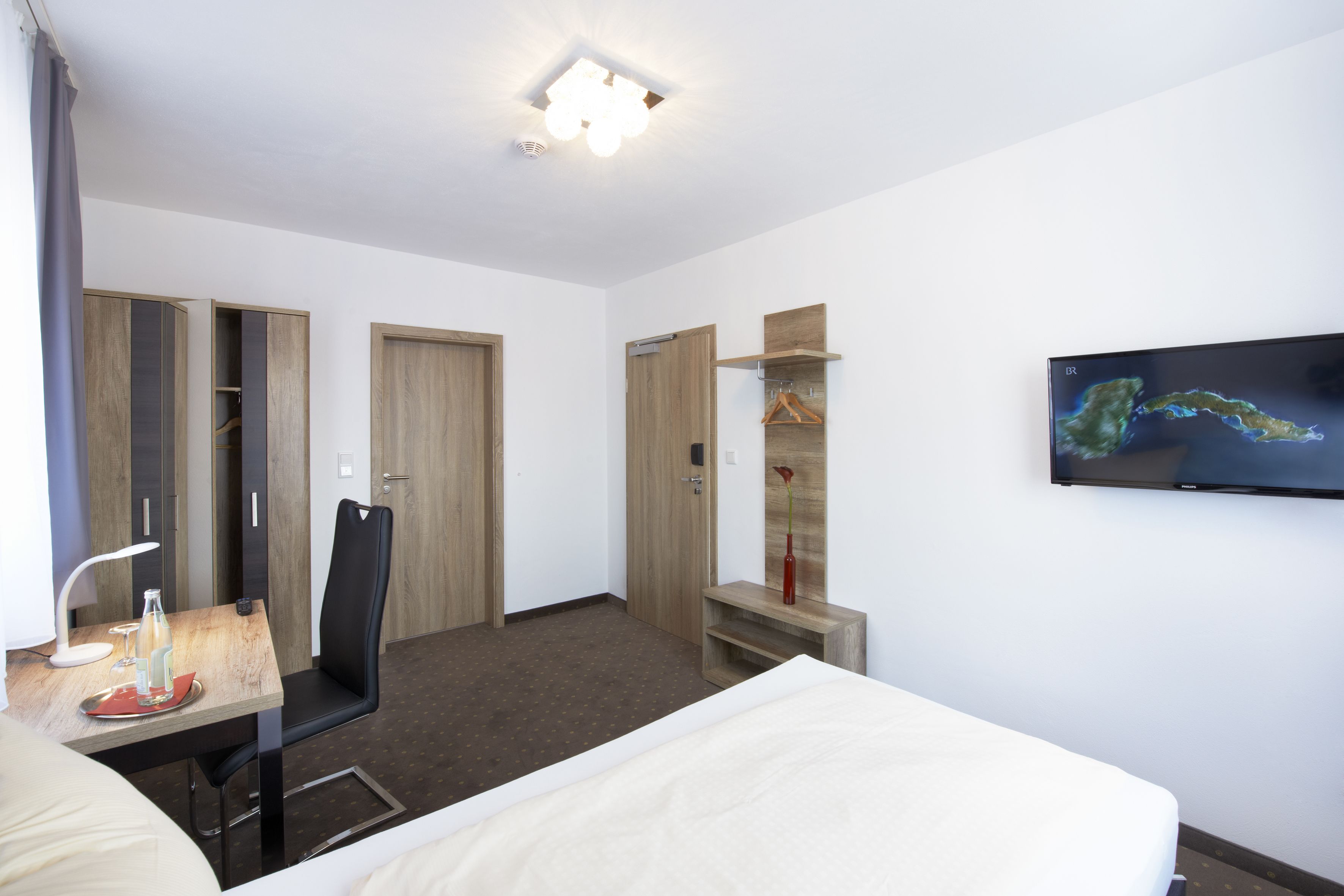 A hotel room with a bed, a desk with a lamp, a chair, a TV mounted on the wall, and a wardrobe.