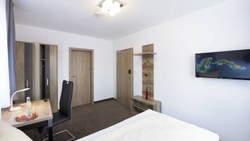 A hotel room with a bed, a desk with a lamp, a chair, a TV mounted on the wall, and a wardrobe.