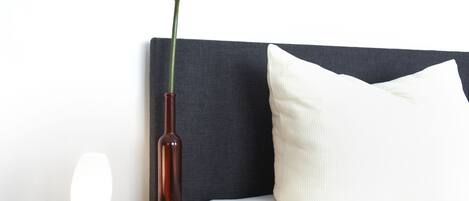 Un lit avec une tête de lit sombre, un oreiller blanc et une table de chevet ornée d’un vase et d’une lampe.