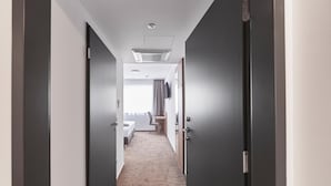 Een hotelgang met donkere deuren, een kamer met tapijtelijke vloerbedekking, een bed, een bureau en een raam met gordijnen.