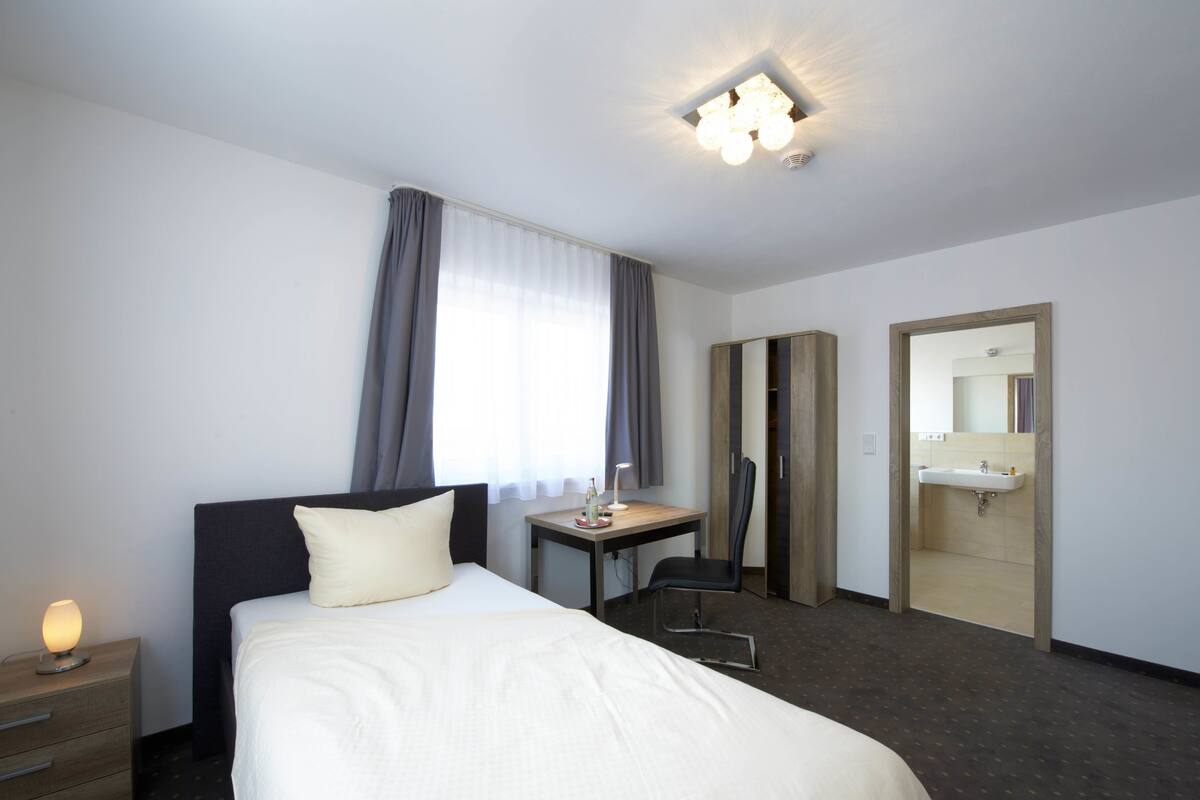 A hotel room with a bed, a desk, a chair, a nightstand, a mirror, and a bathroom.