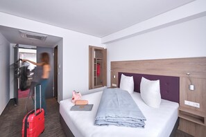A hotel room with a bed, a wooden headboard, a bedside table, a wardrobe, and a suitcase. - HOTEL NEW IN - Ingolstadt - Gaimersheim (Gaimersheim)