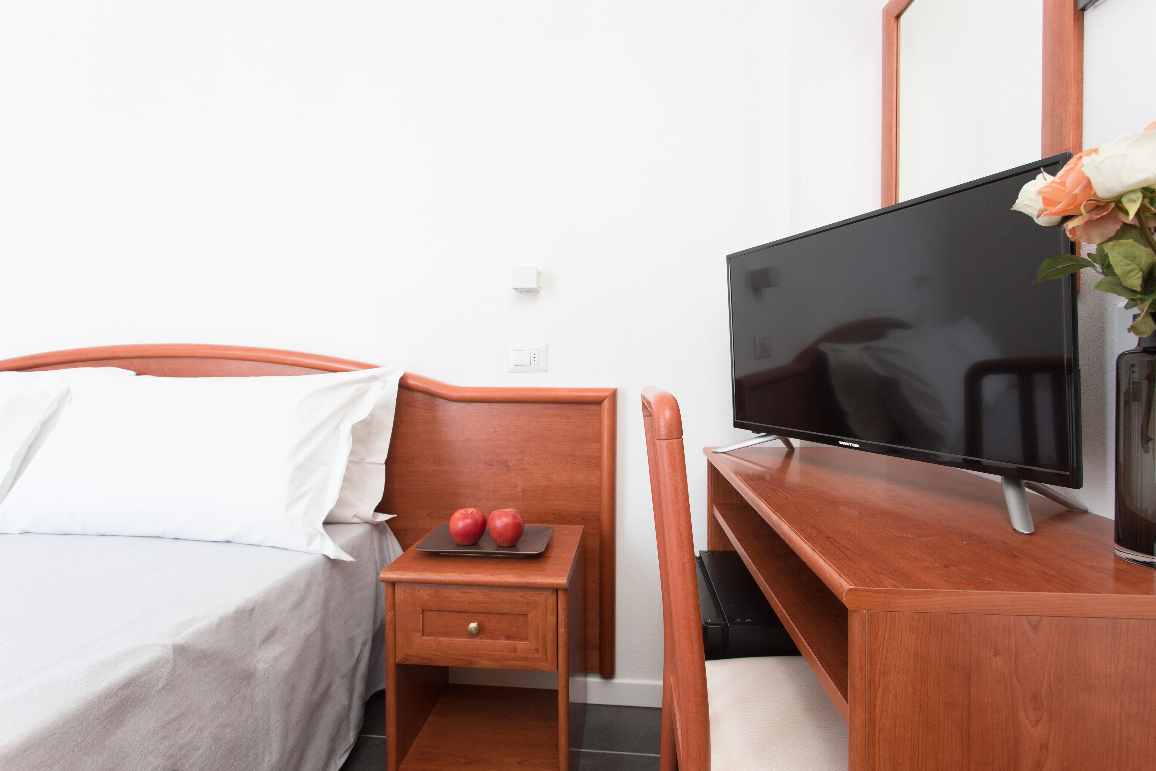 Habitación de hotel con una cama, una mesita de noche de madera con una bandeja que contiene dos manzanas, un televisor sobre un mueble de madera y un florero con flores.