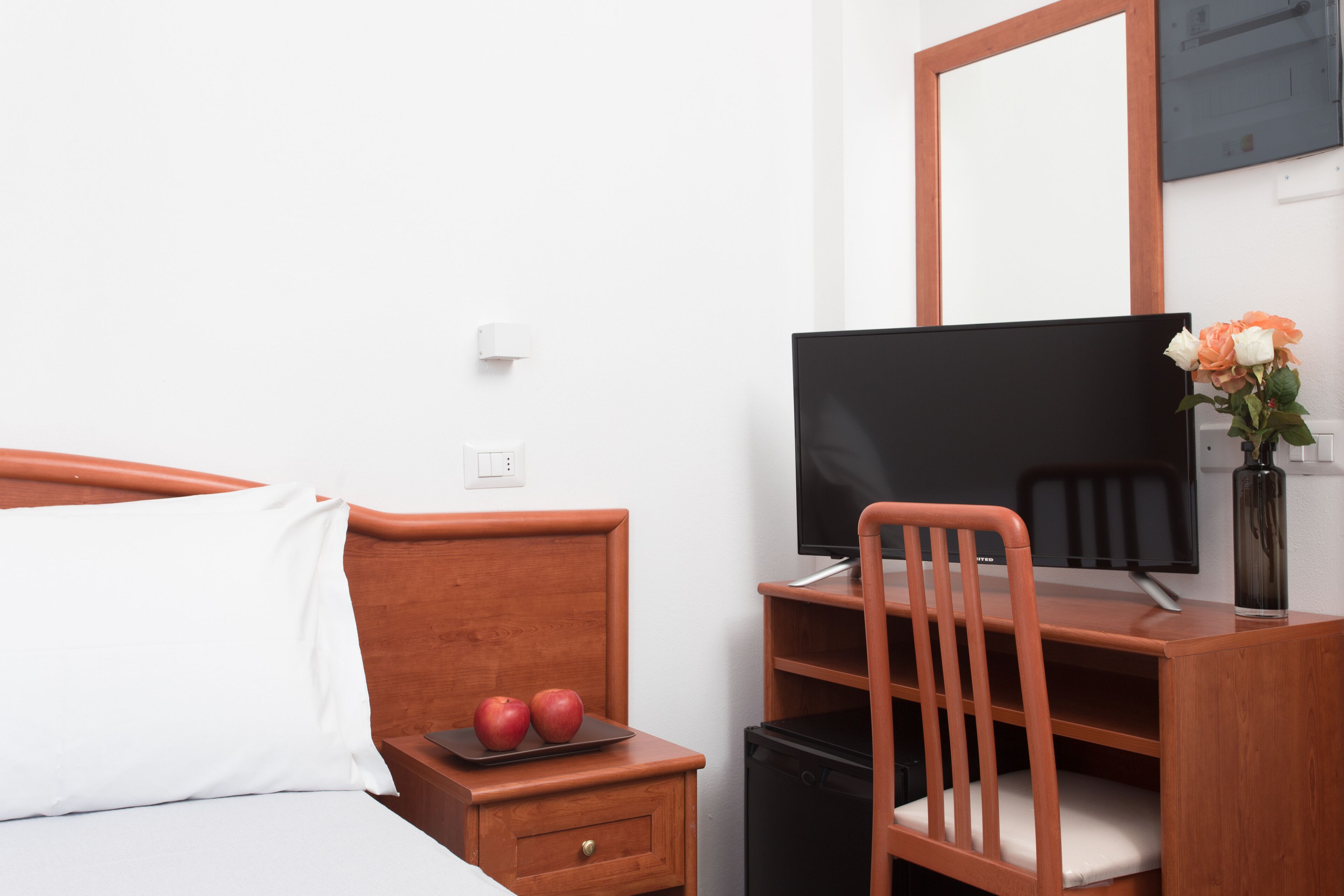 Ett hotellrum med en säng, ett träbord vid sängen med två äpplen, ett skrivbord med en stol, en tv och en vas med blommor.
