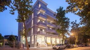 Un hotel de varios pisos con balcones, una entrada visible y un estacionamiento con coches.