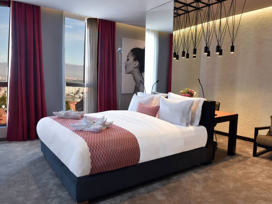 A modern hotel room with a large bed, red curtains, a city view, and a unique ceiling light fixture.