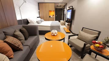 A modern hotel room with a sofa, two round orange tables, a chair, a TV, and a fruit basket.