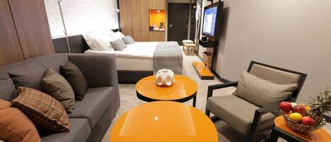 A modern hotel room with a sofa, two round orange tables, a chair, a TV, and a fruit basket.