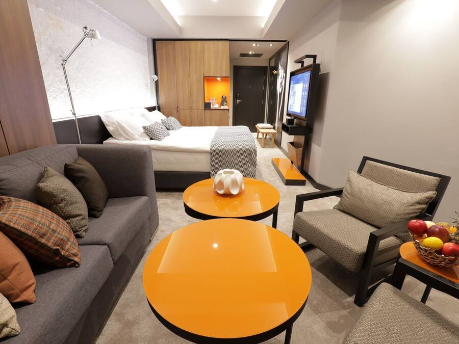 A modern hotel room with a sofa, two round orange tables, a chair, a TV, and a fruit basket.