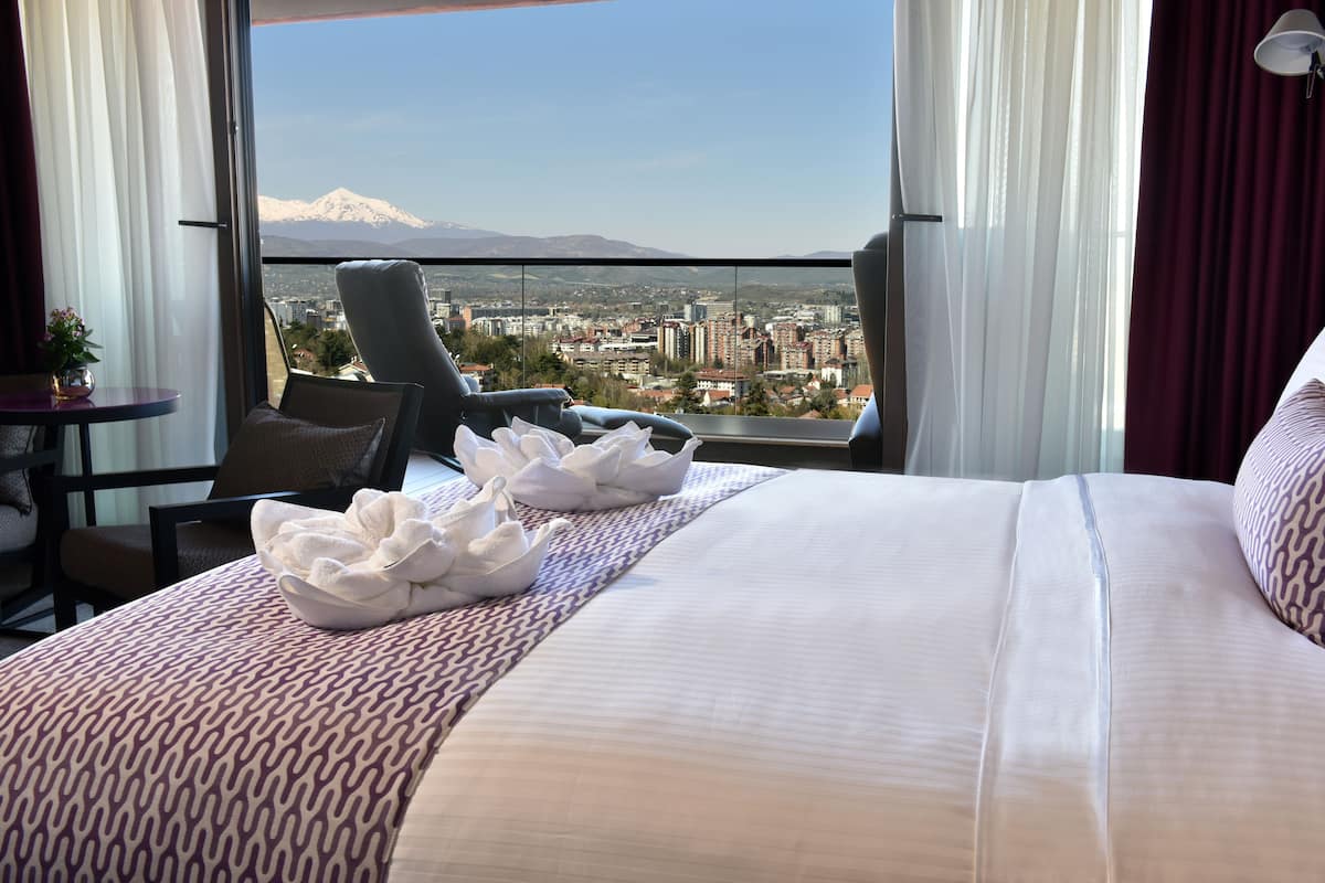 Habitación de hotel con una cama grande, una silla, una mesita y vistas a la ciudad y montañas.