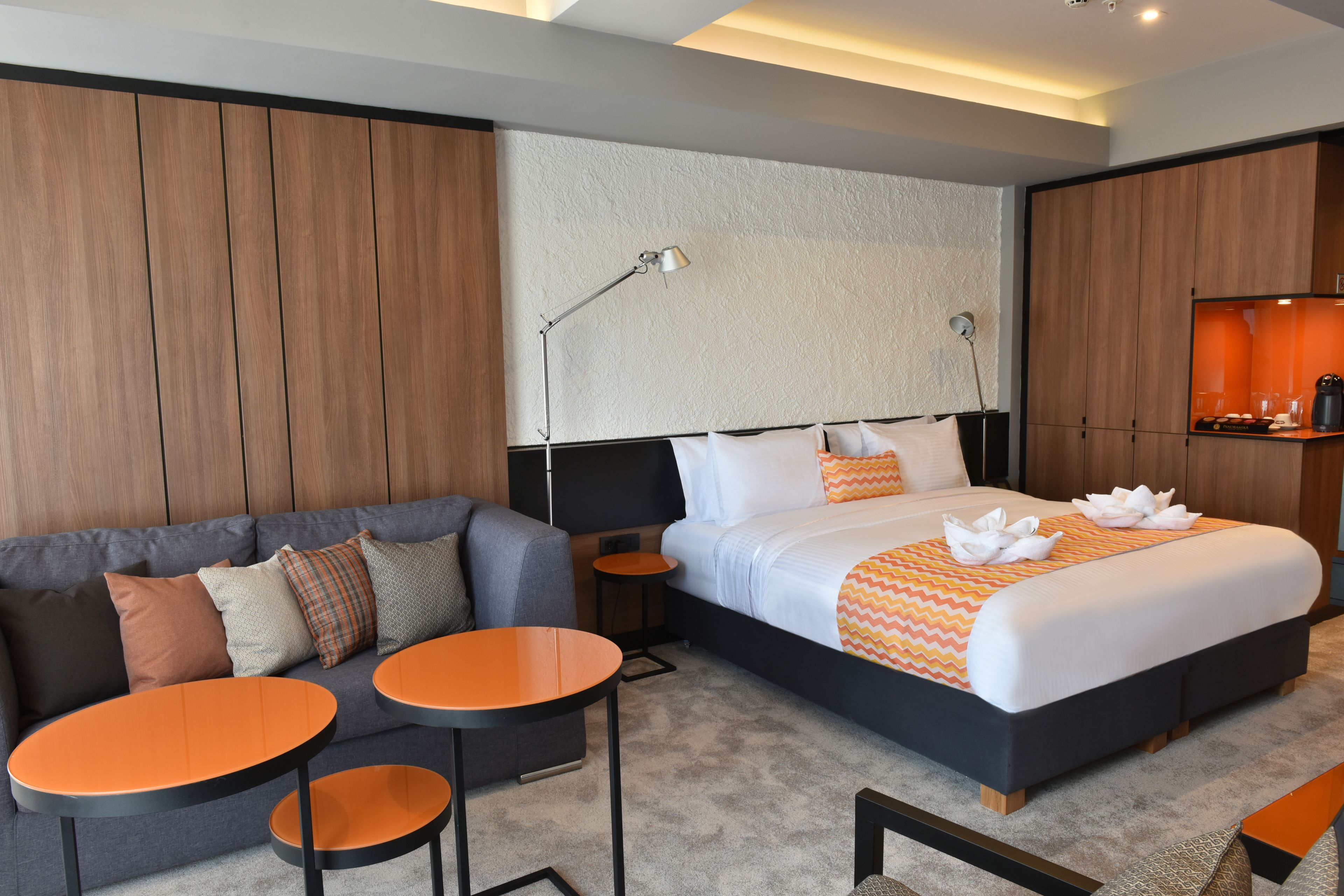 Una habitación de hotel moderna con una cama grande, un sofá, dos mesitas pequeñas y un televisor montado en la pared.