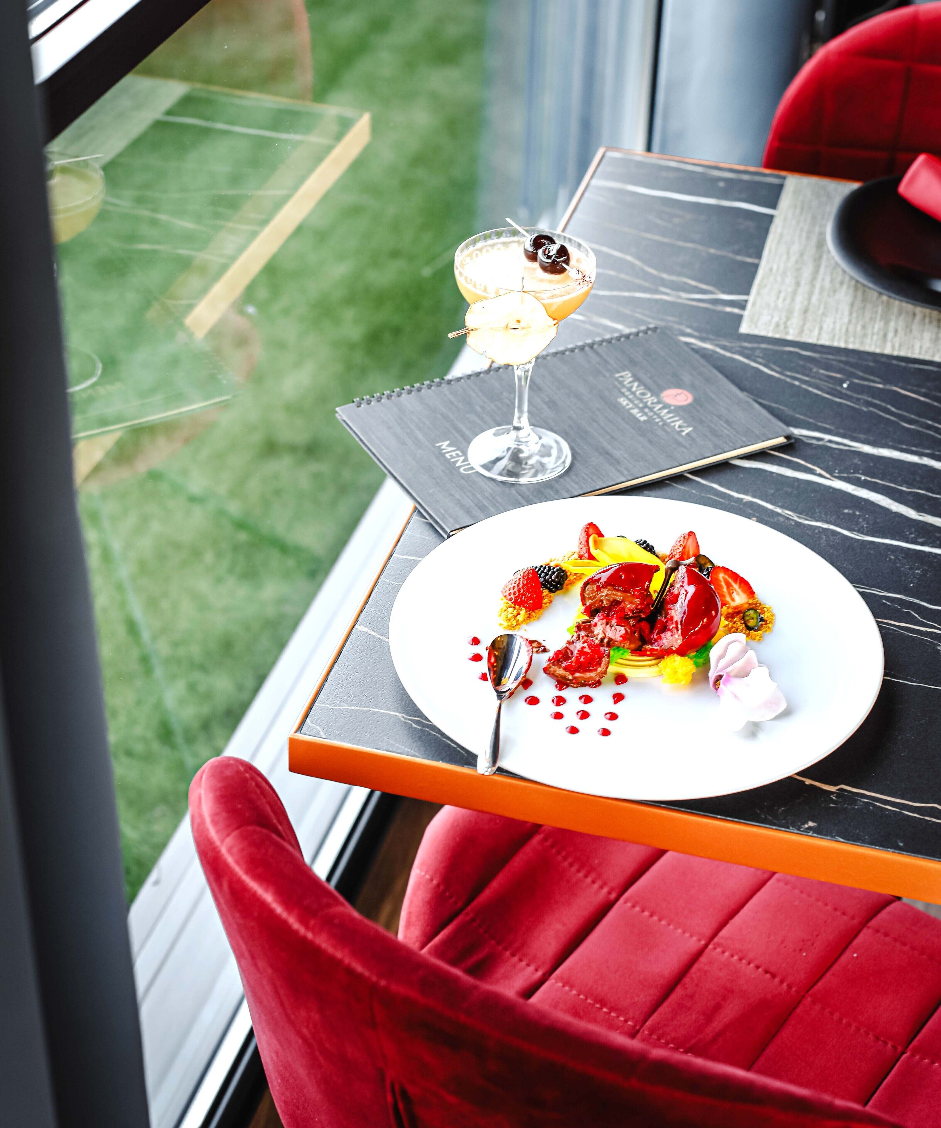 Ein Tisch mit Dessert und Speisekarte, rote Stühle und Blick auf eine Rasenfläche.