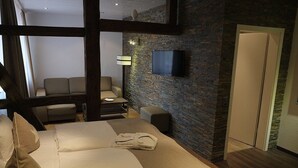 Una habitación de hotel con una cama grande, una zona de estar con sofá y sillones, un televisor de pantalla plana montado en una pared de ladrillo, y una puerta que da a otra habitación.