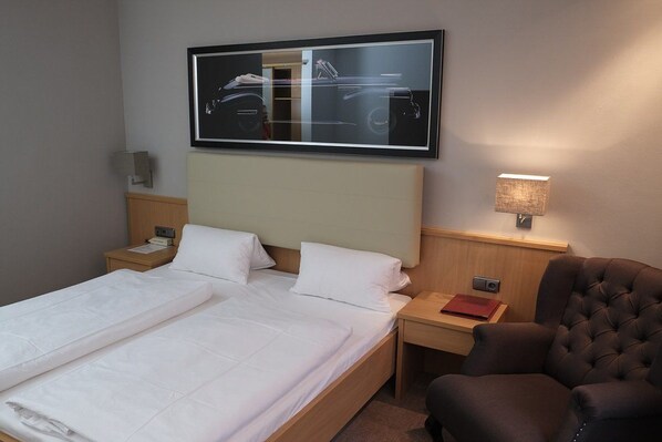 A hotel room with a large bed, a headboard, a nightstand with a lamp, a chair, and a framed picture on the wall. - Hotel Brunnenhof International (Wedemark)