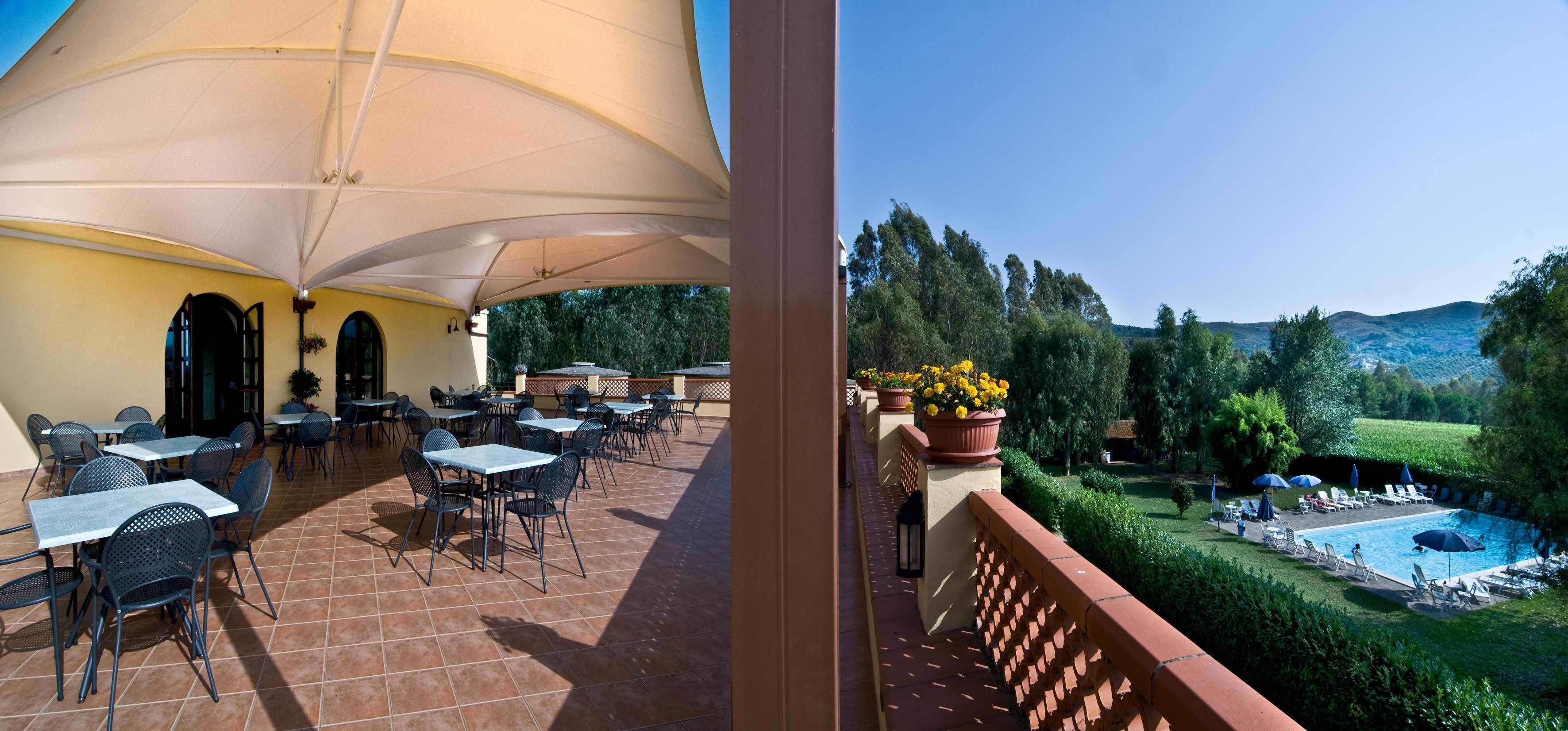 An outdoor terrace with tables and chairs, a pool area with sun loungers, and a view of the surrounding landscape.