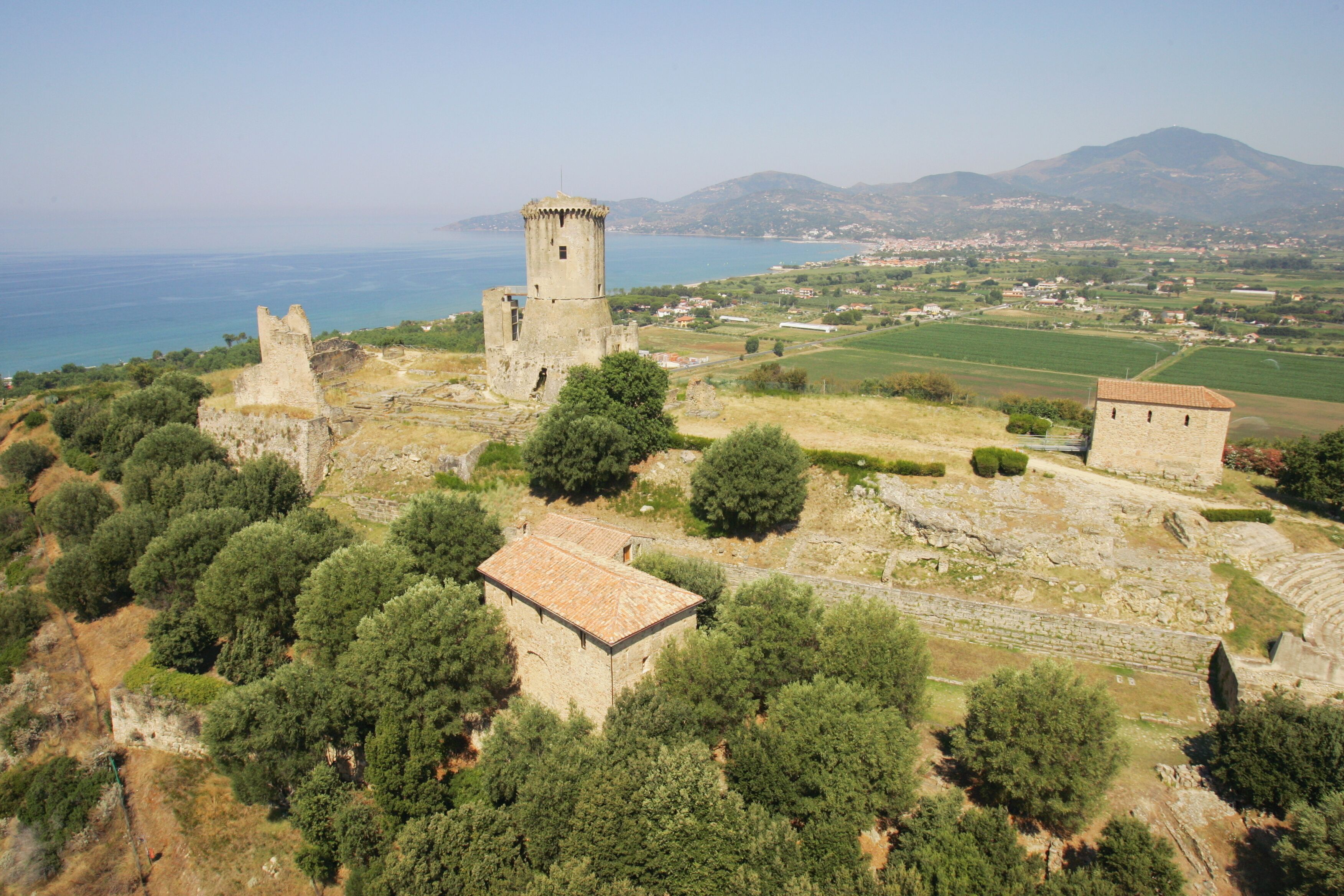 Vue aérienne d’un site historique en ruines, comprenant une tour et un bâtiment à toiture de tuiles, le tout entouré d’une végétation luxuriante et avec un paysage côtier en arrière-plan.