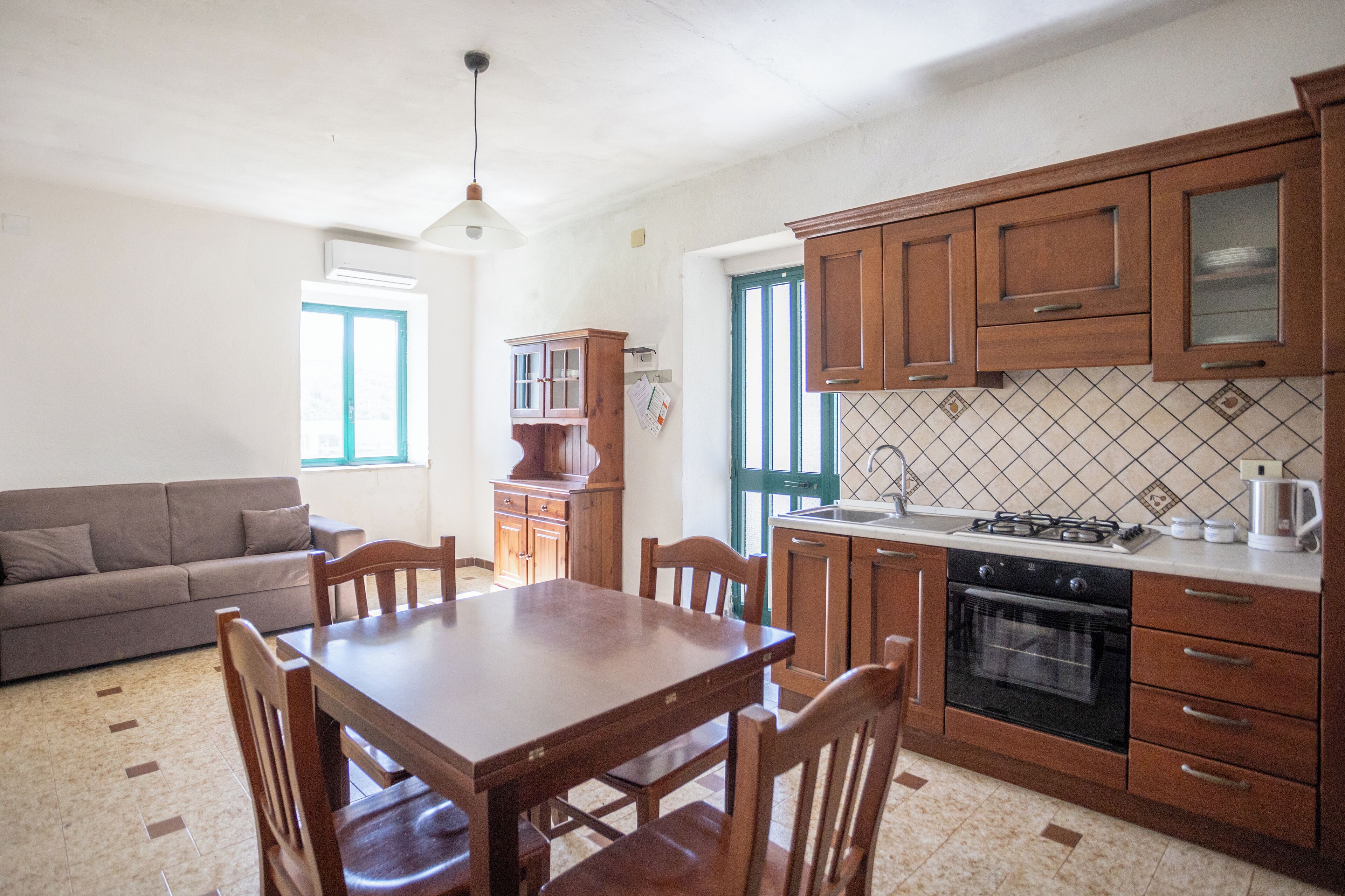 Une cuisine avec des meubles de rangement en bois, une table à manger, des chaises, un canapé et une fenêtre.